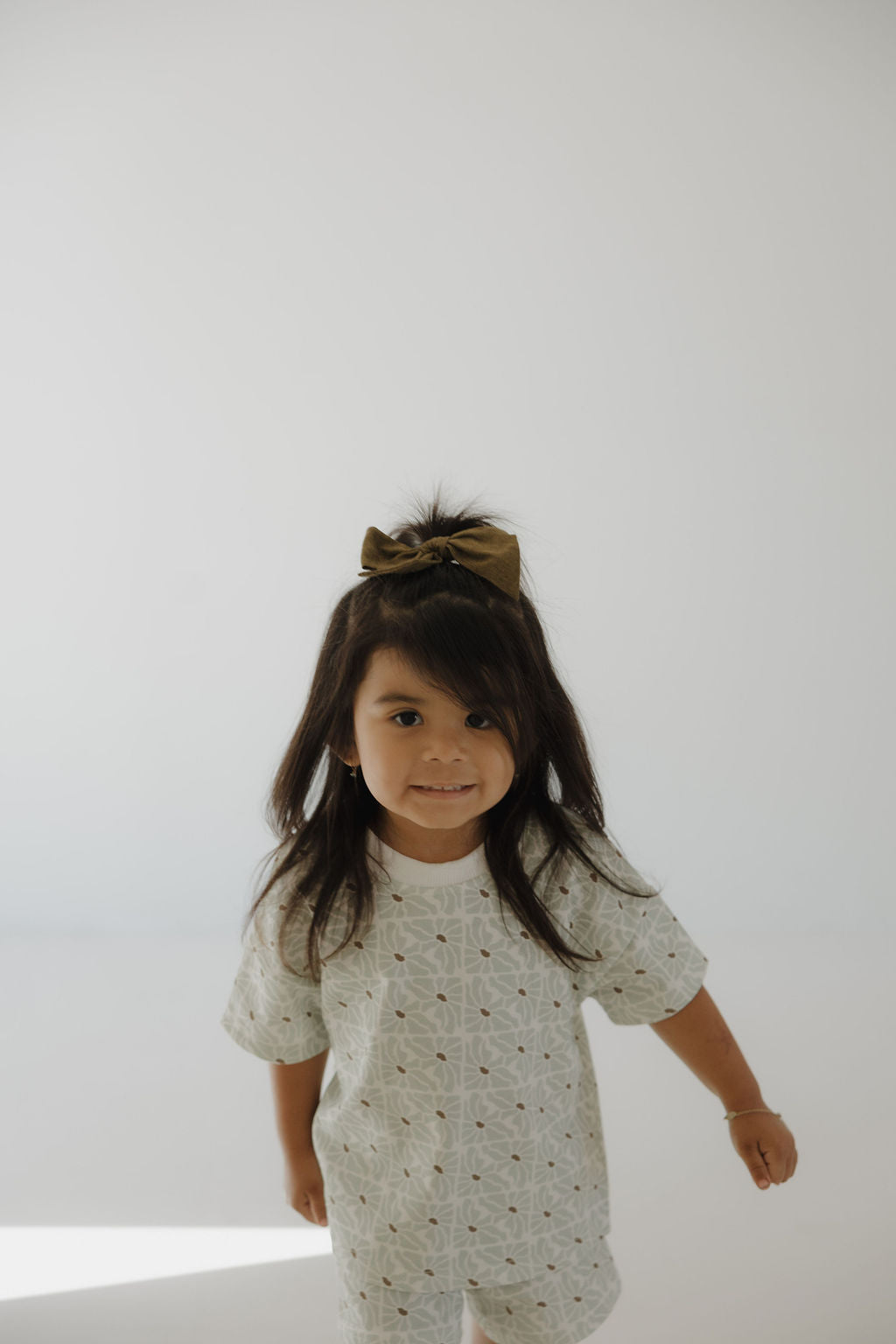 A young girl with long dark hair and a large bow smiles gently at the camera in a bright, minimalistic room, wearing the forever french baby French Terry Short Set | Coast.