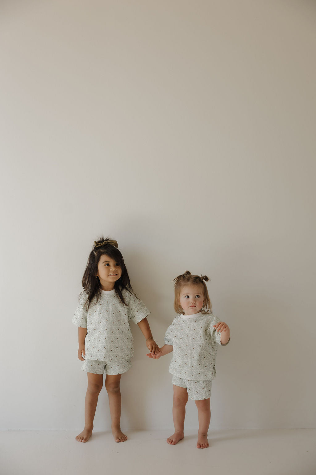Two barefoot children stand against a plain wall, holding hands and wearing the forever french baby French Terry Short Set in Coast. Their matching outfits feature subtle patterns and an FF logo, styled with small top knots in their hair.