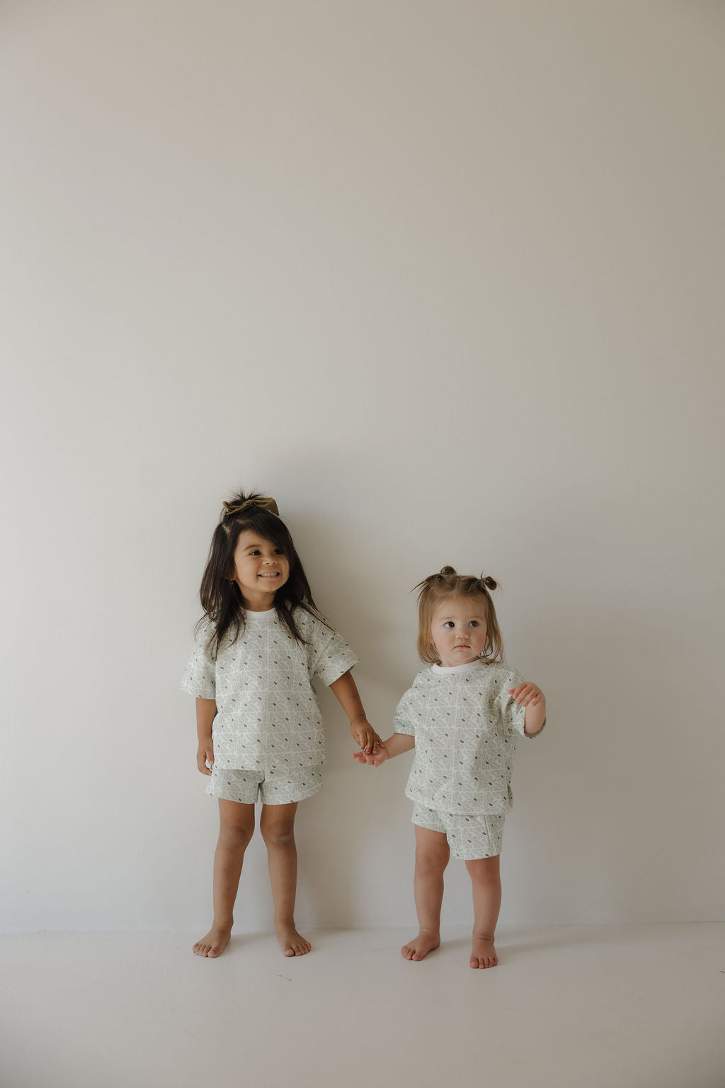 Two young children stand barefoot against a plain wall, holding hands. Both wear the forever french baby French Terry Short Set in Coast, featuring matching shorts and a soft short sleeve top with an FF logo detail.