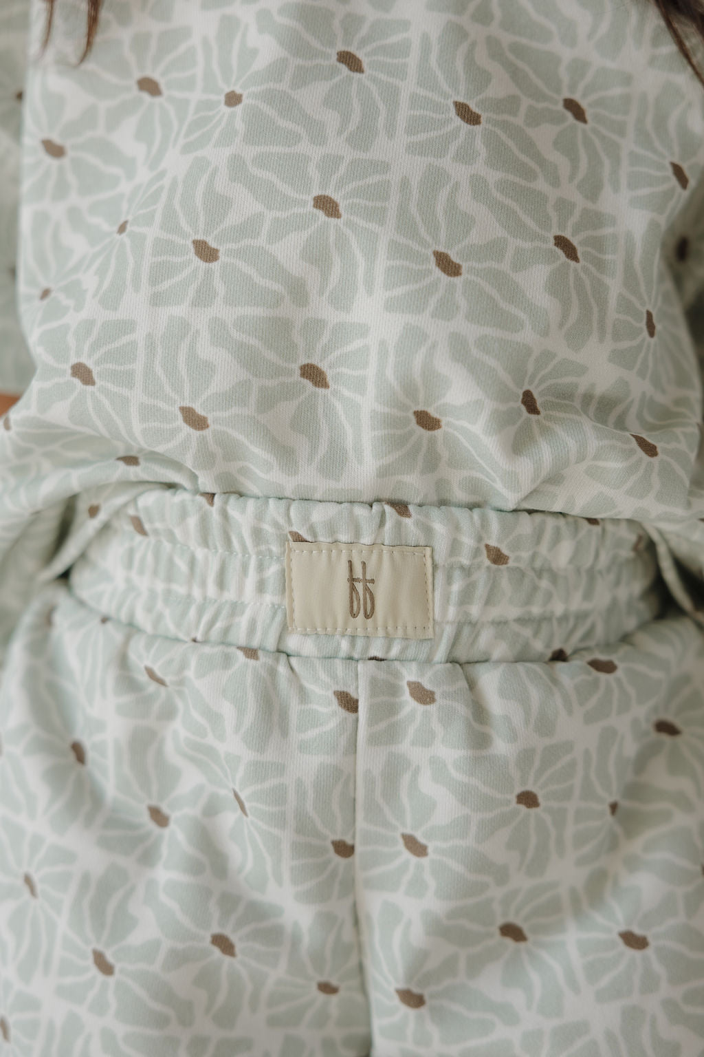 Close-up of a person wearing the French Terry Short Set | Coast from forever french baby, featuring a light-colored short sleeve top and matching shorts with a white and brown leaf pattern and a beige "bb" tag on the waistband.