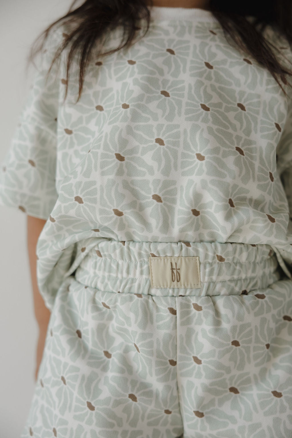 A child wears the forever french baby French Terry Short Set in Coast, featuring light green shorts and a short sleeve top with a subtle leaf and dot pattern. The elastic waistband sports a small beige "bb" tag. The child's face is not visible.