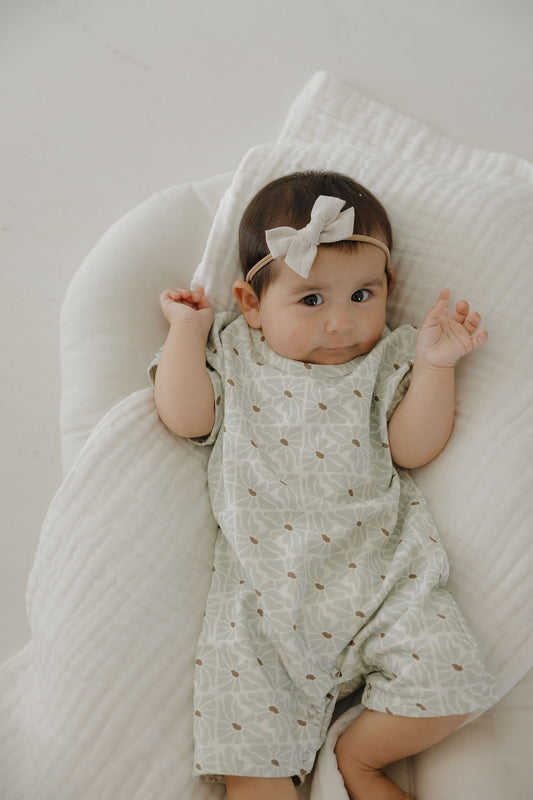 A baby with dark hair wearing the forever french baby French Terry Romper | Coast and a white bow headband lies on a soft white blanket, looking up with raised hands and a calm expression.