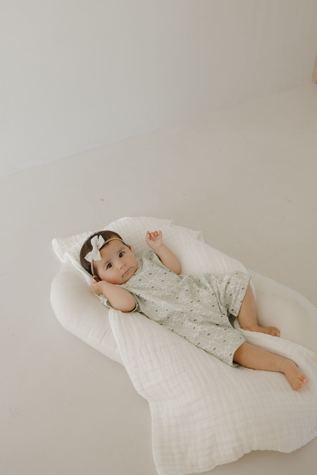 A baby in a forever french baby French Terry Romper | Coast and a white bow headband lies on a soft, white cushion, one arm raised. The minimalistic, light-toned background highlights the cozy romper.