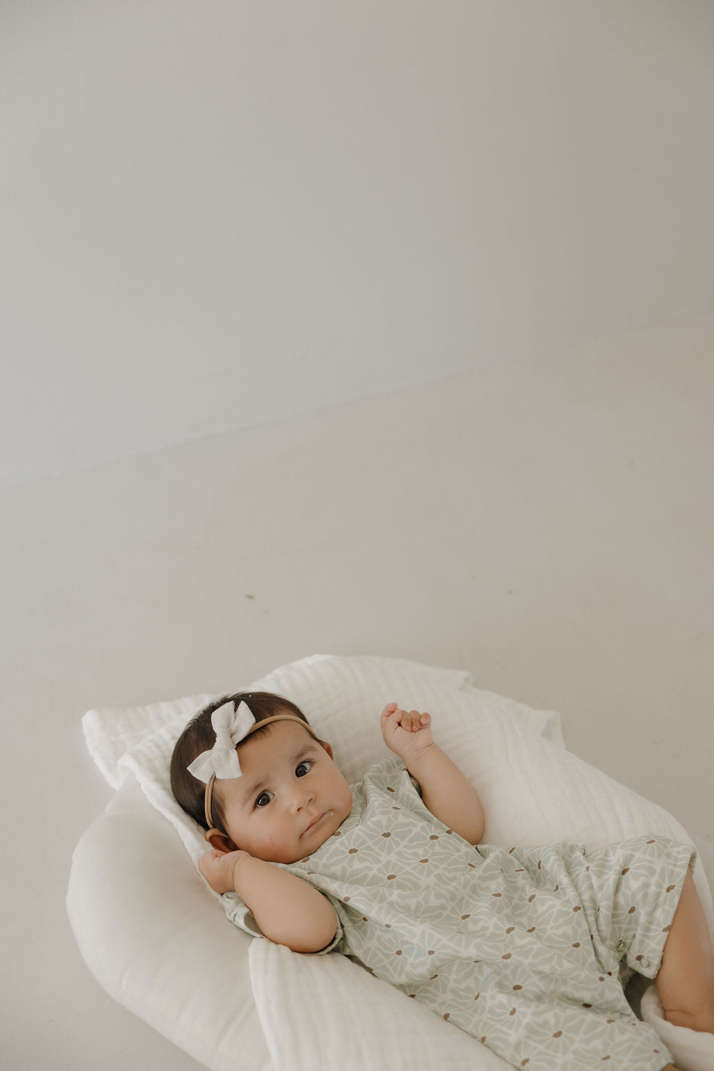 A baby in the forever french baby French Terry Romper | Coast, a light green patterned and breathable newborn outfit, paired with a white bow headband, lies on a soft white cushion against a plain background, looking up with one arm raised.