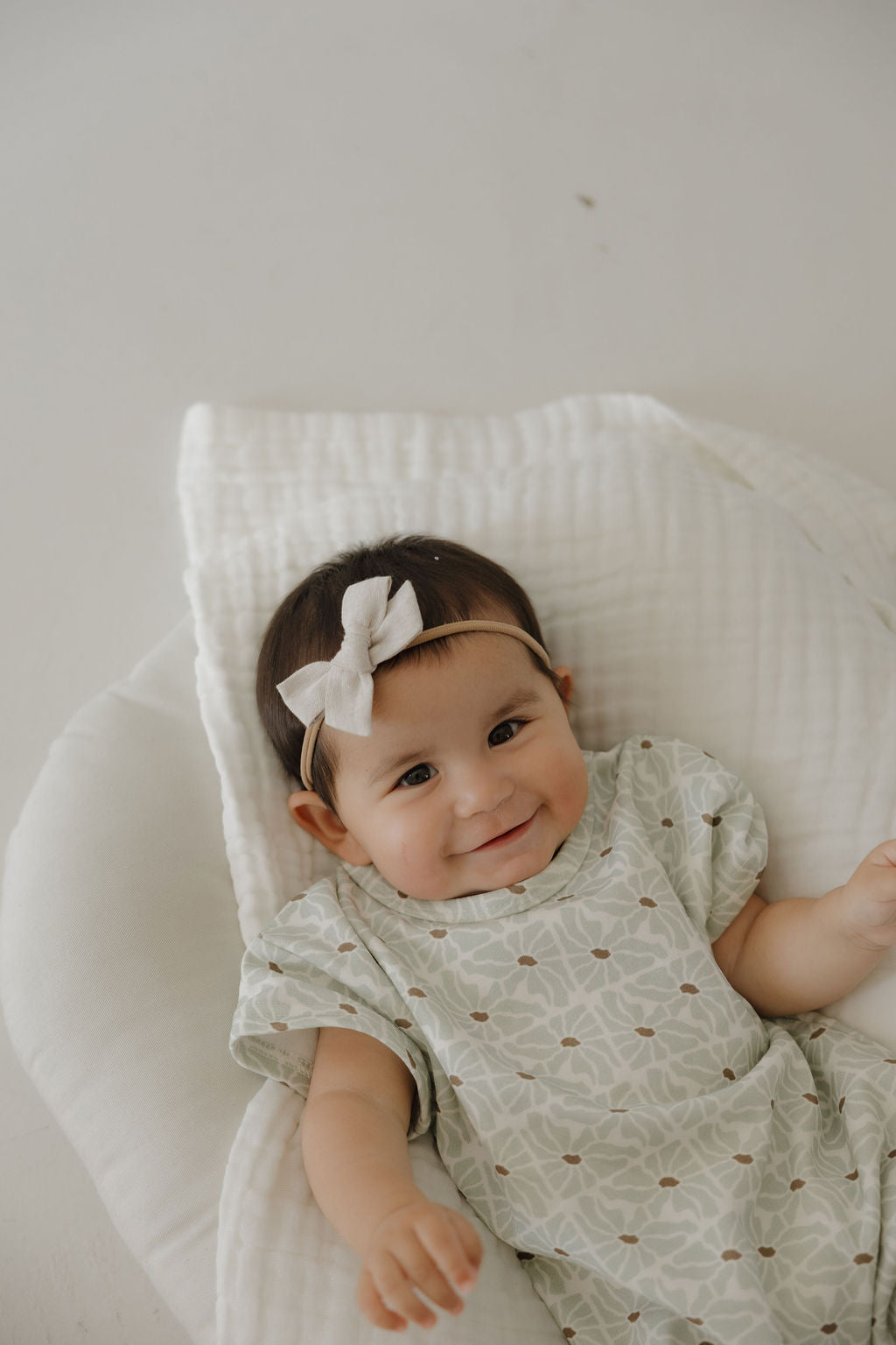 A smiling baby in the forever french baby French Terry Romper | Coast lies on a white cushioned blanket, wearing a light-colored bow headband and looking up at the camera.