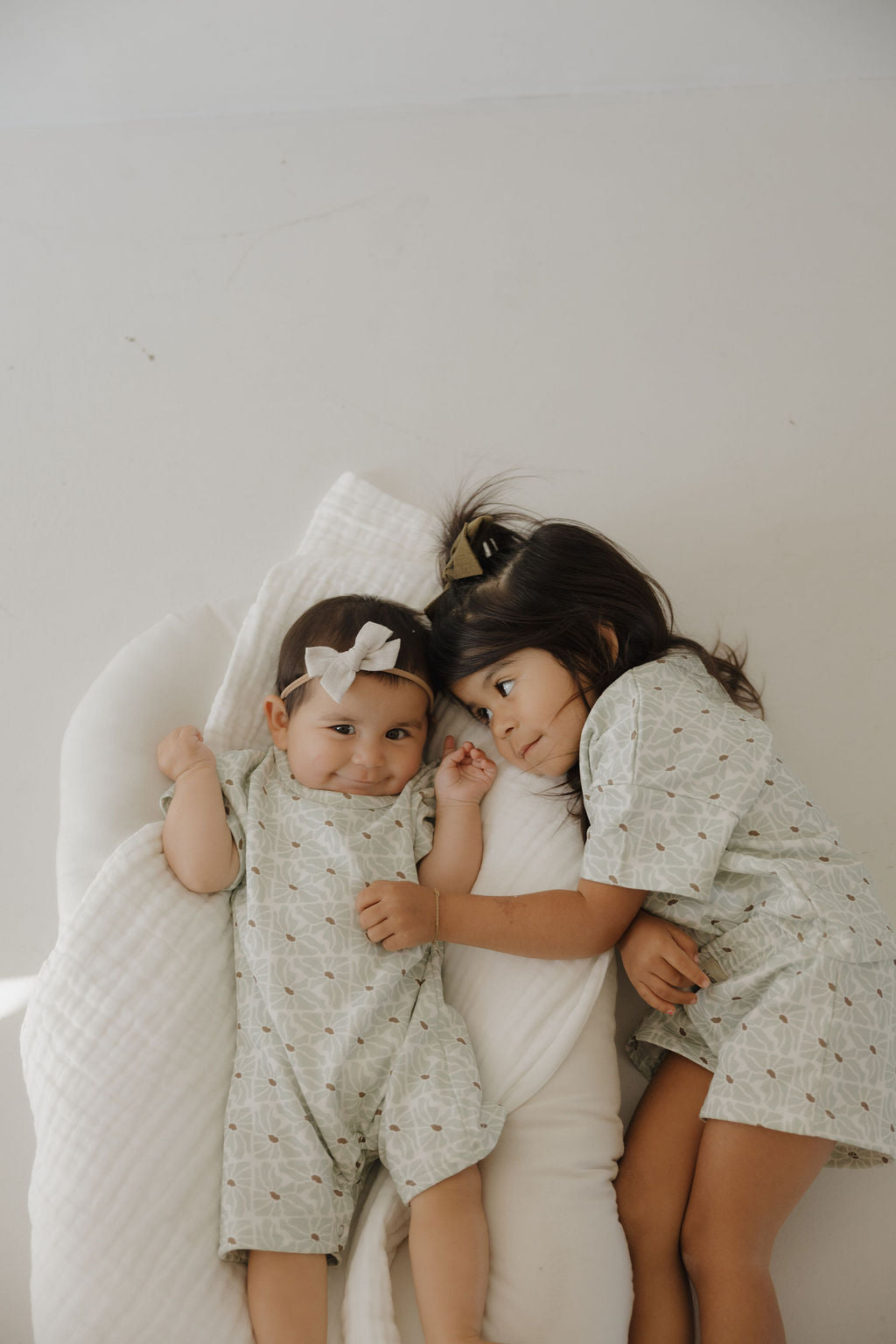 Two young children lie close together on a white pillow. The baby, dressed in the forever french baby French Terry Romper | Coast and a bow headband, is gently touched by the older child. Both look calm and affectionate.