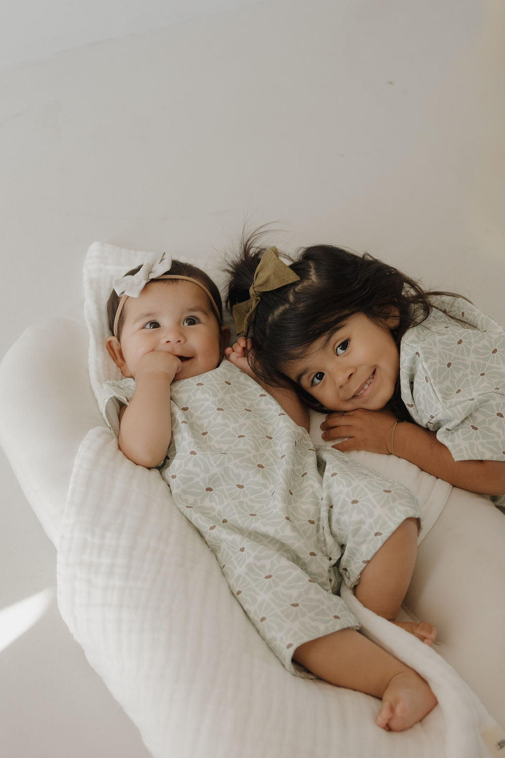 Two young children, a baby and toddler, snuggle on a soft white blanket wearing matching forever french baby French Terry Rompers in Coast. The geometric patterned outfits are light green, breathable, and paired with hair bows as they smile at the camera.