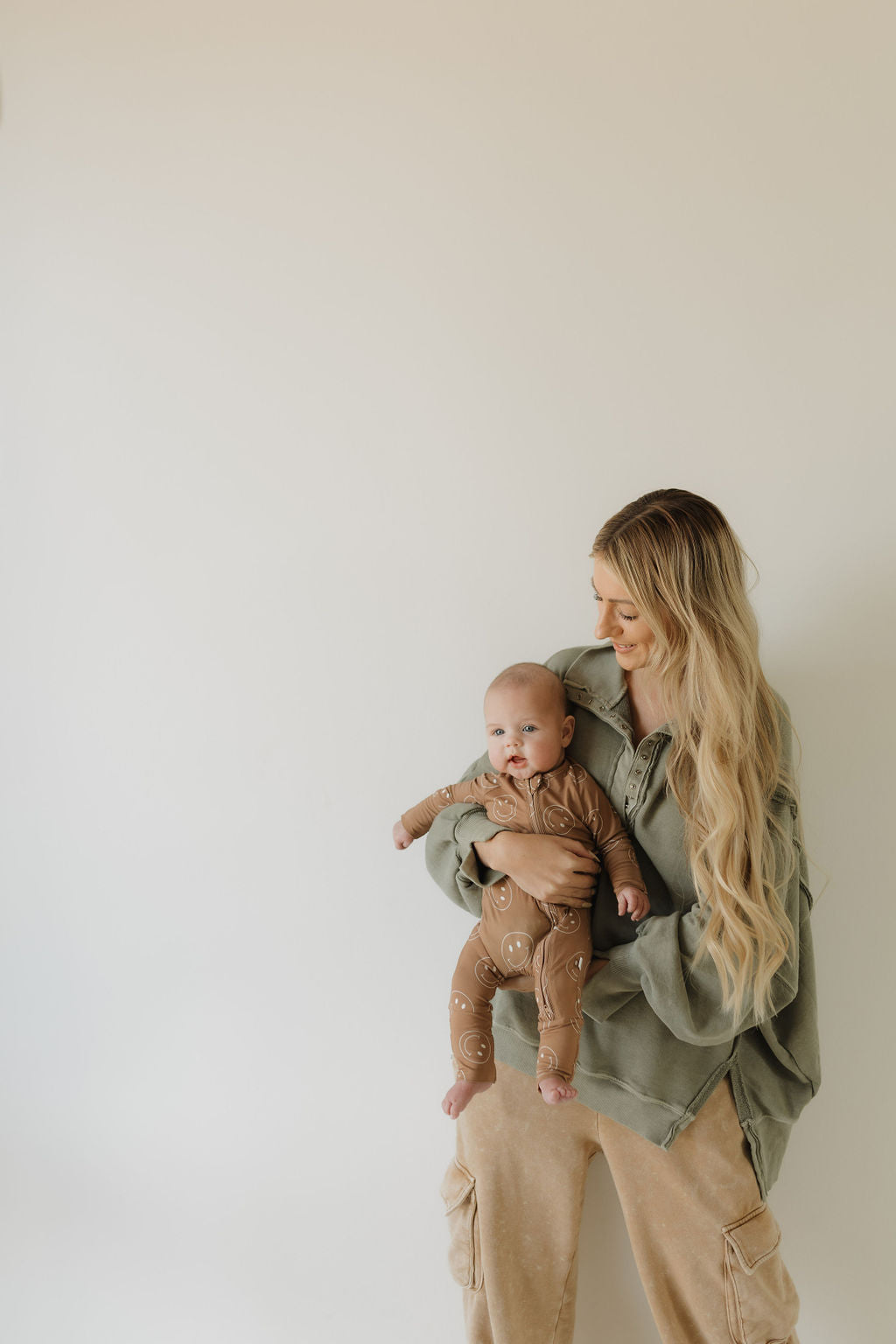 A woman with long blonde hair cradles a baby dressed in forever french baby's Bamboo Zip Pajamas in Sandstone Smiles. She wears a green jacket and beige pants, and they lovingly gaze at each other against a plain white background, embodying comfort and warmth.