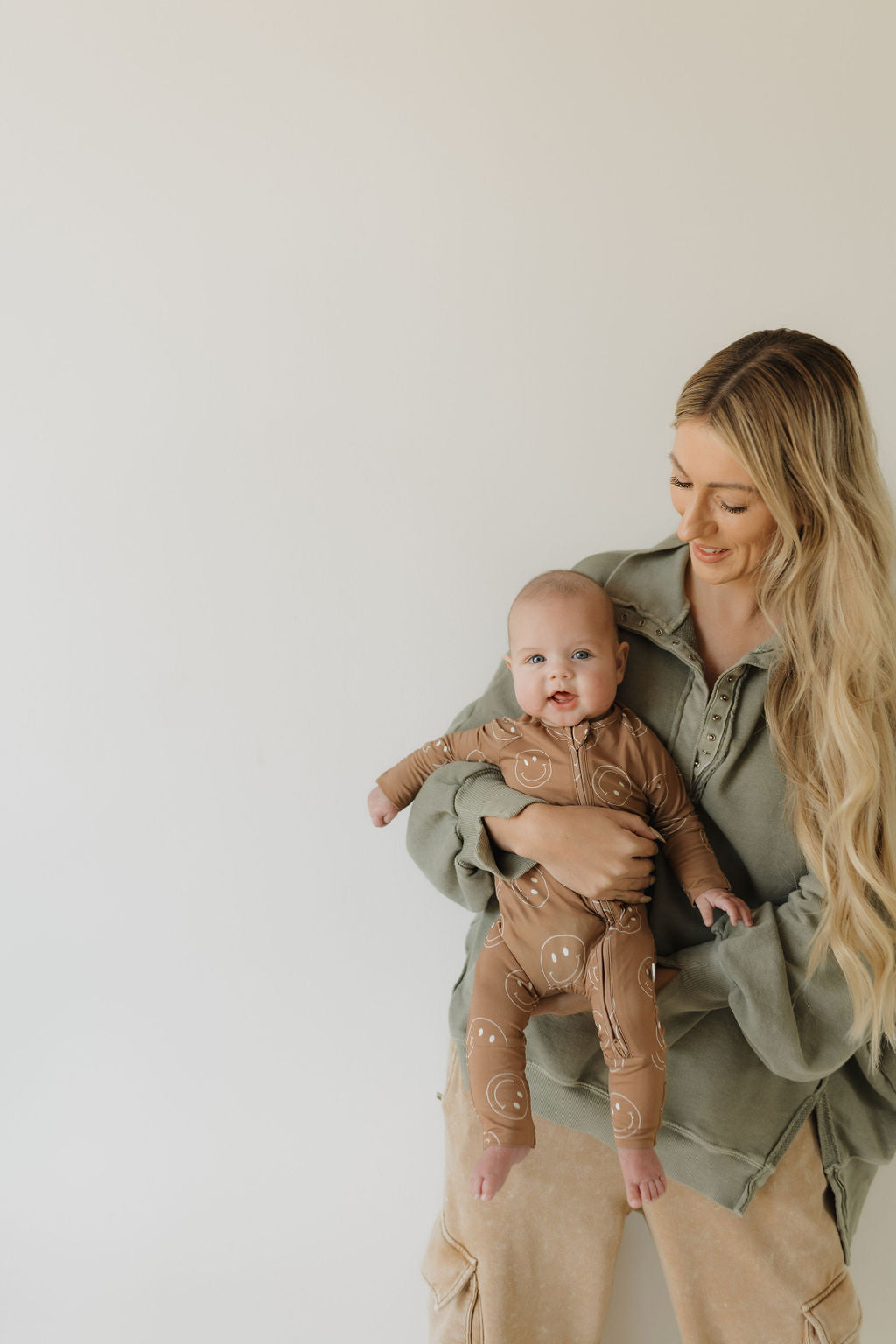 A woman with long blonde hair smiles while holding a baby in Bamboo Zip Pajamas, "Sandstone Smiles," by forever french baby. She stands against a plain light background in an oversized green sweater and beige pants, exuding warmth and comfort.