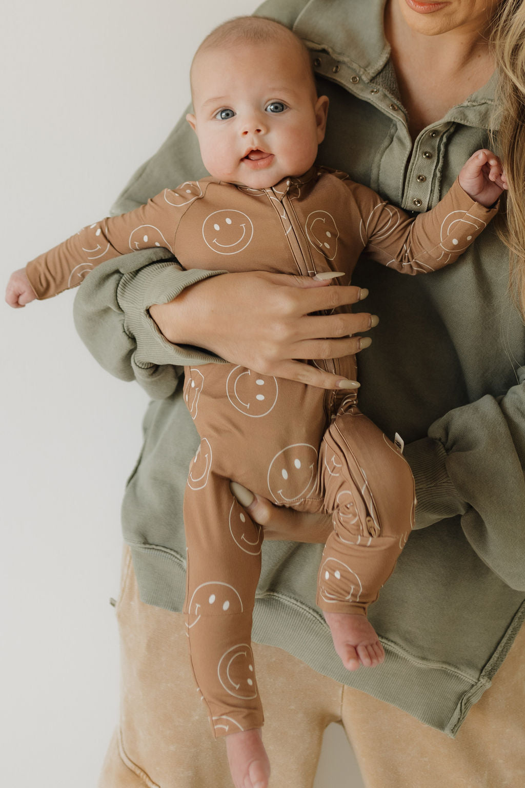 A baby in a Bamboo Zip Pajamas | Sandstone Smiles by forever french baby, featuring smiley faces on breathable material, is held by someone in a green sweatshirt. The little one gazes up with curiosity.