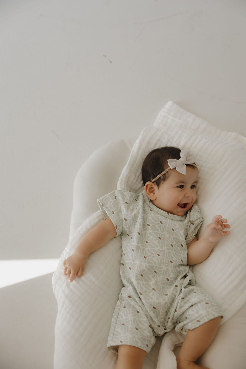 A smiling baby in the forever french baby French Terry Romper | Coast lies on a soft, white cushion with a light blanket and a bow headband, gazing off to the side.