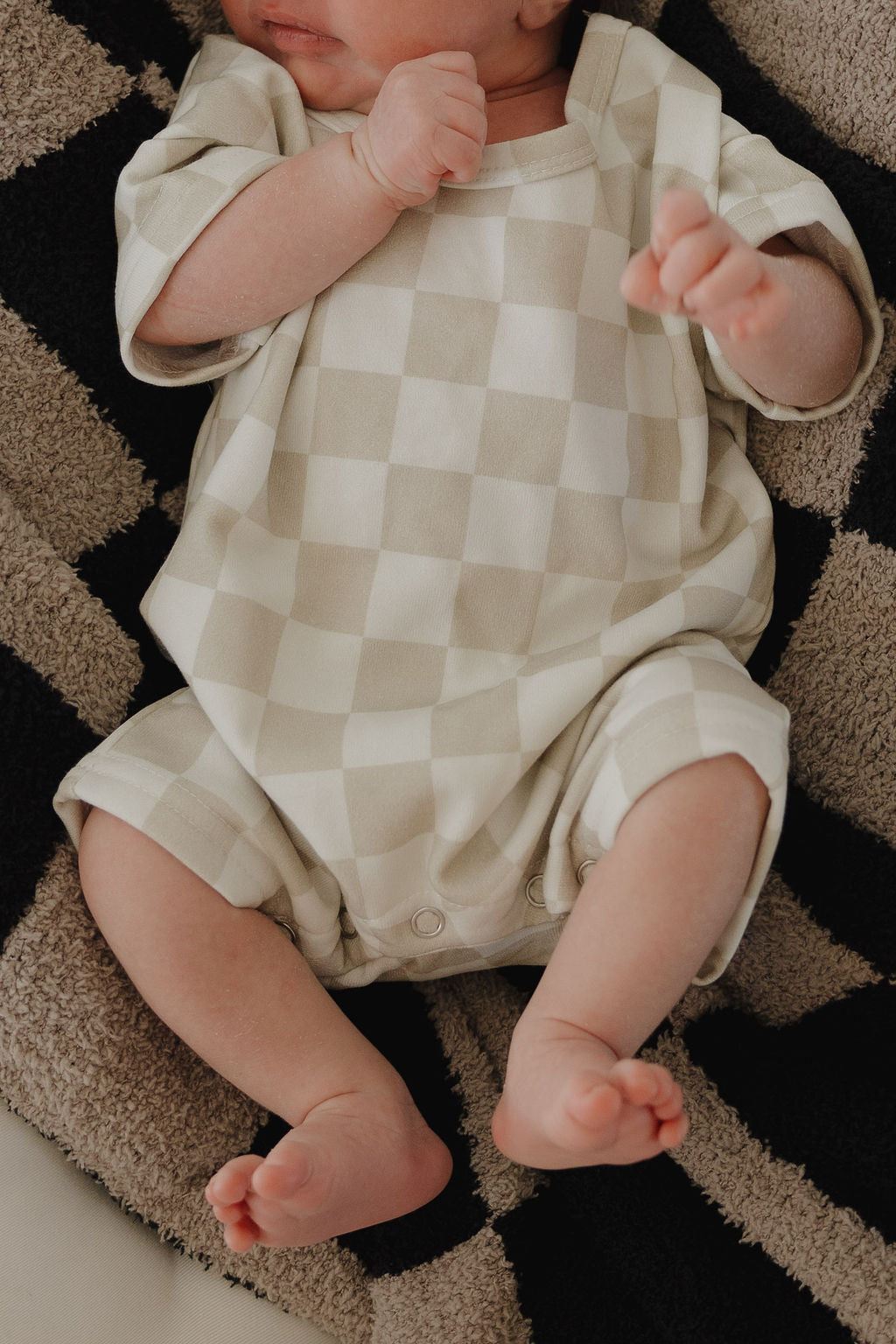A baby in the forever french baby French Terry Romper | Checkerboard lies on a striped textured blanket, arms bent and legs apart. The beige and white checkered romper features convenient snap closures. The baby's face is partially out of frame.