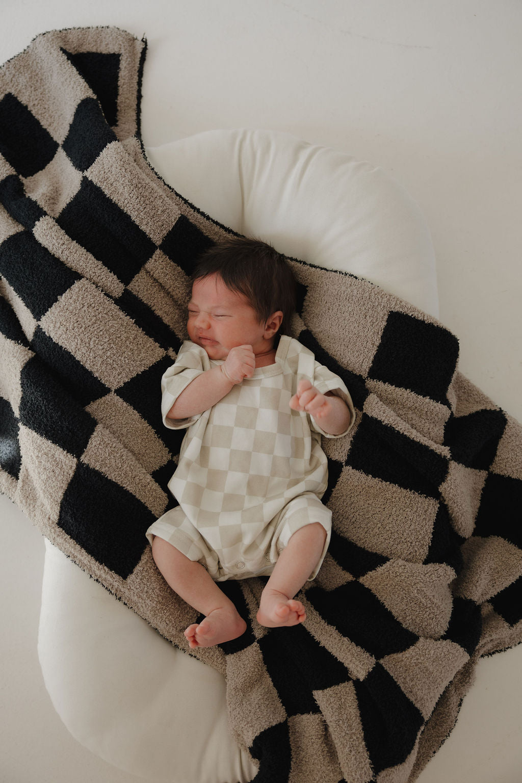 A newborn sleeps on a white cushion, wrapped in a black and beige checkered blanket and wearing the forever french baby French Terry Romper | Checkerboard, all bathed in soft lighting against a minimal background.