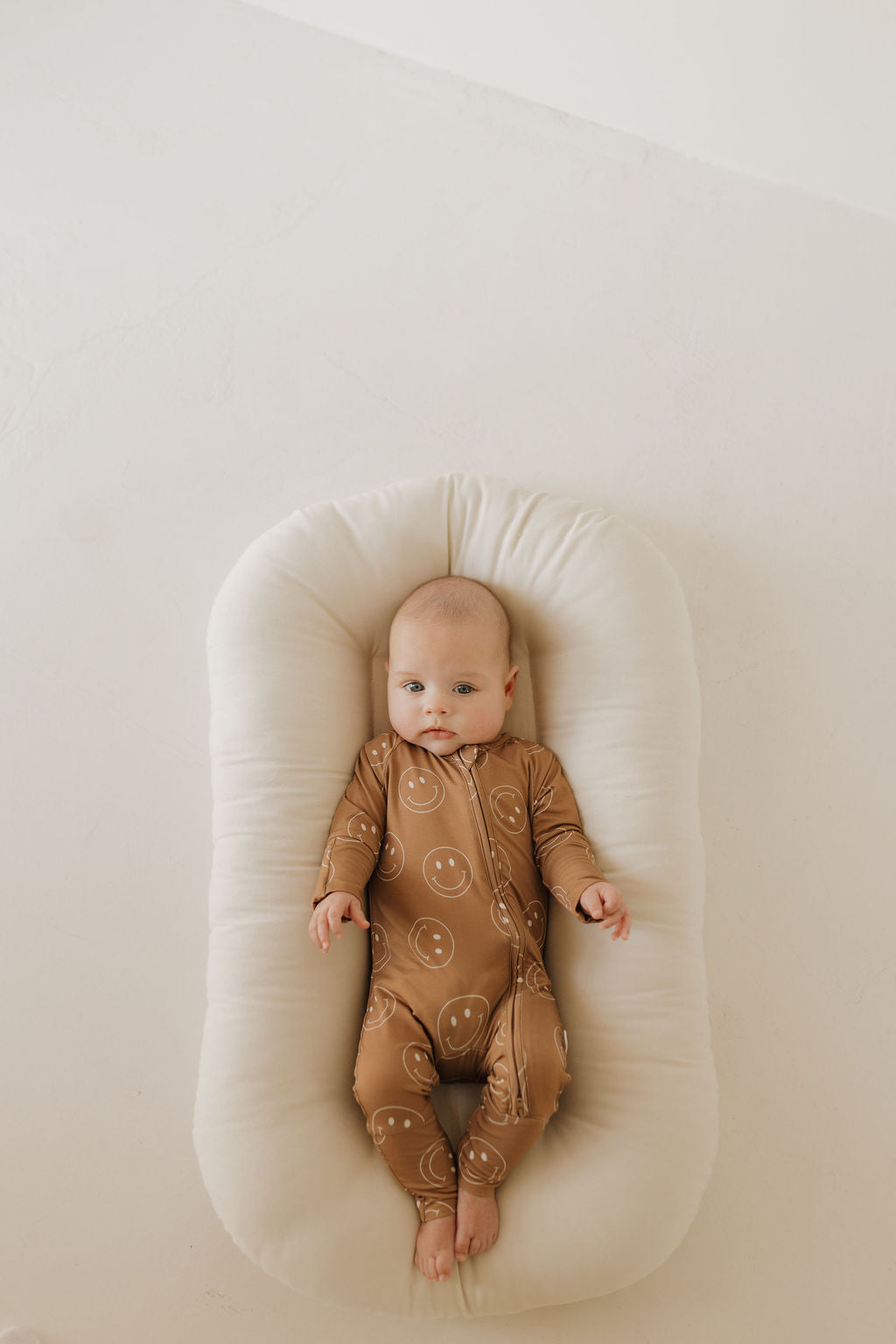 A baby wearing forever french baby's Bamboo Zip Pajamas in Sandstone Smiles rests on a cushioned white lounger. The hypo-allergenic attire, alongside the light cream background, ensures comfort and style.