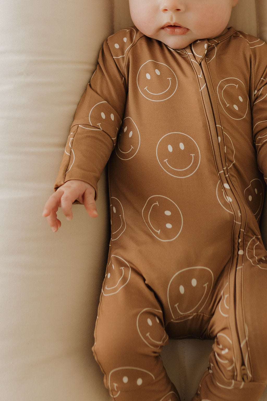 A baby in "forever french baby" Bamboo Zip Pajamas | Sandstone Smiles is shown with focus on their torso and legs, one hand slightly outstretched. Hypo-allergenic made from breathable fabric with white smiley patterns on brown, set against a soft neutral-toned background enhancing coziness.