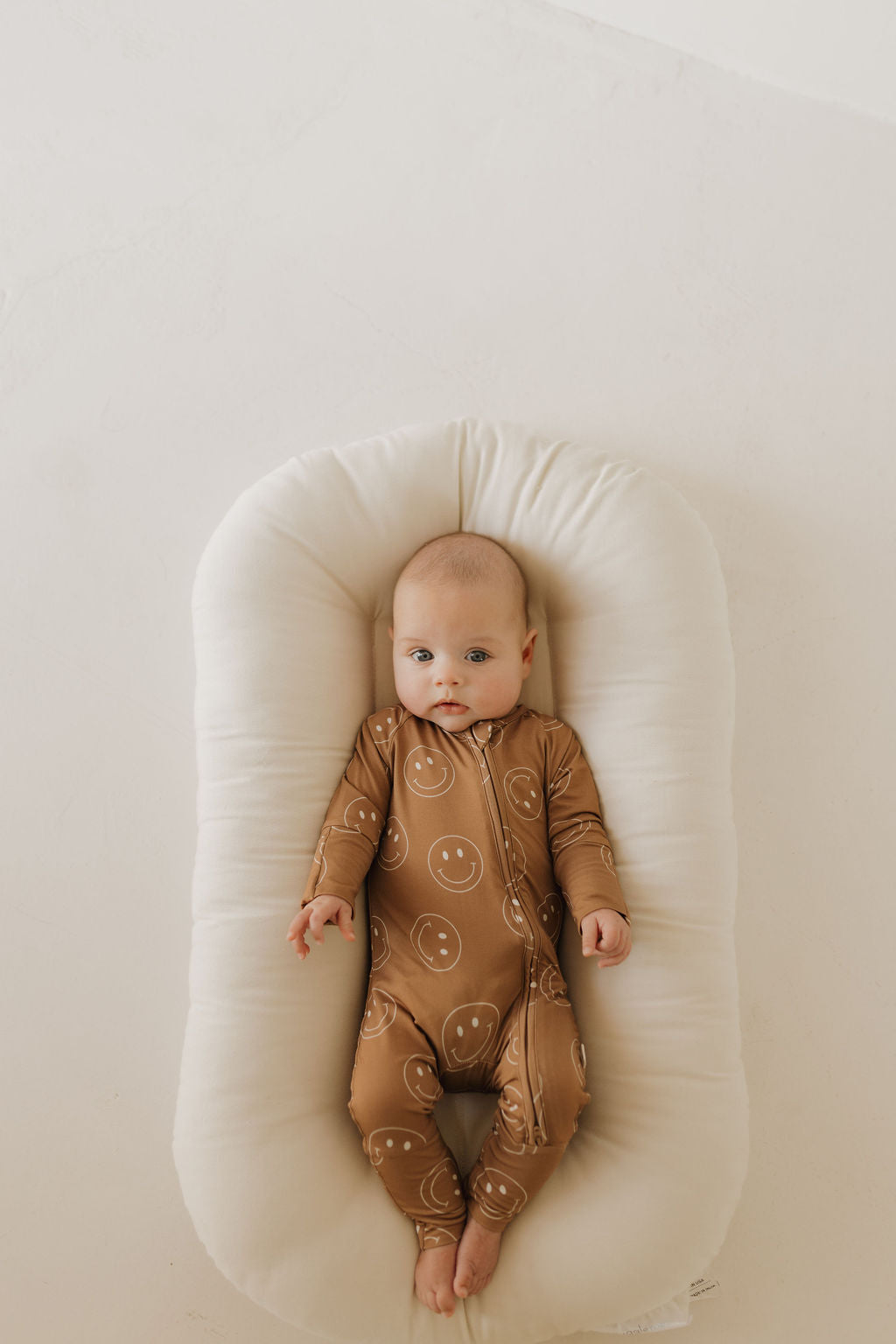 A baby in a Bamboo Zip Pajamas | Sandstone Smiles by forever french baby lies on a white cushioned surface, gazing upwards. The breathable sleepwear ensures comfort in the softly toned room.