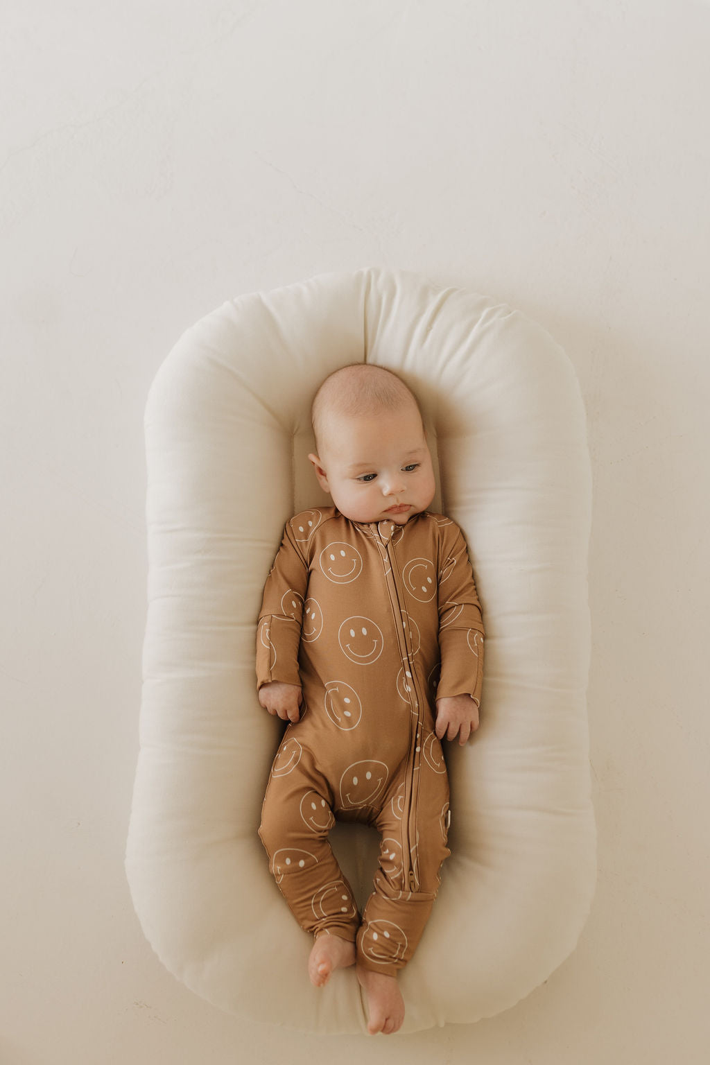 A baby in "Sandstone Smiles" bamboo zip pajamas by forever french baby lies on a soft, white lounger with a calm expression, gazing to the side, against a neutral light background.