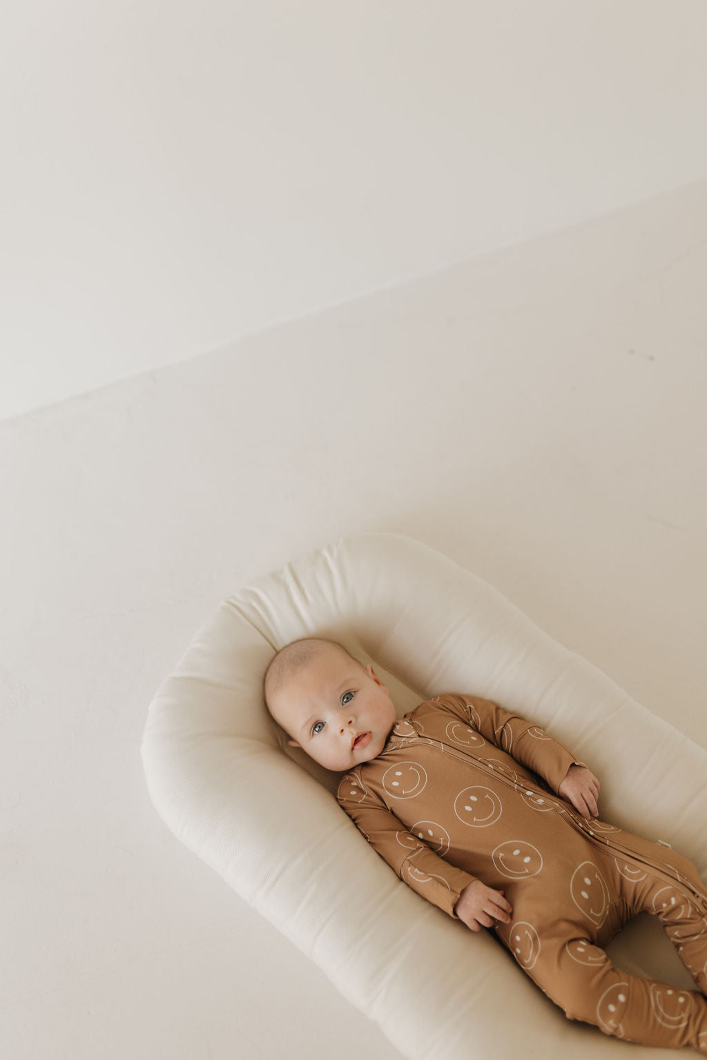 A baby rests on a soft, white cushion dressed in breathable Bamboo Zip Pajamas from forever french baby featuring the Sandstone Smiles pattern. The light and minimalistic background enhances this serene scene.