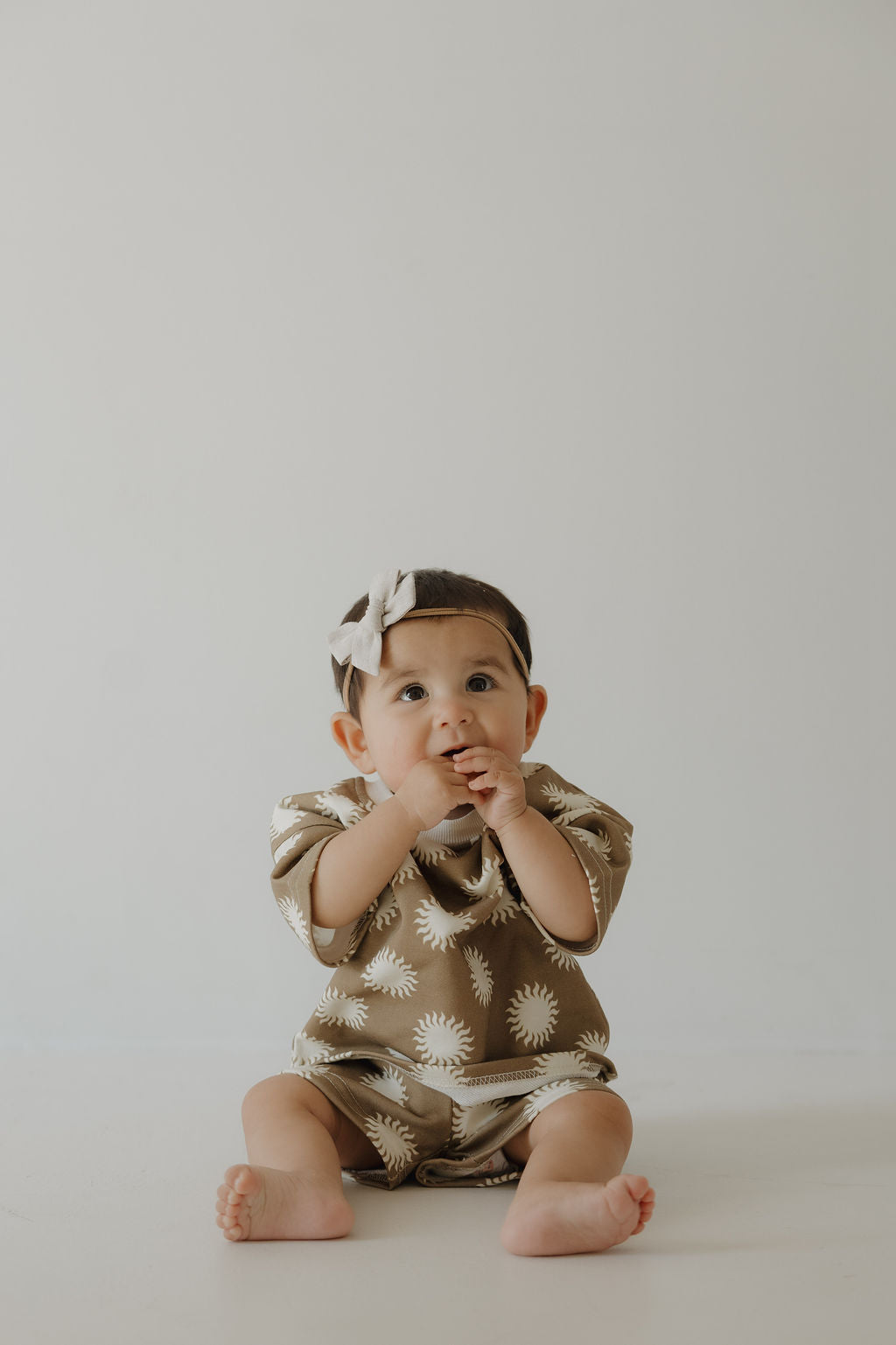 A baby in the French Terry Short Set | Sol by forever french baby—brown with white sunburst, matching shorts and headband—sits on a light background, looking upward with hands near the mouth.