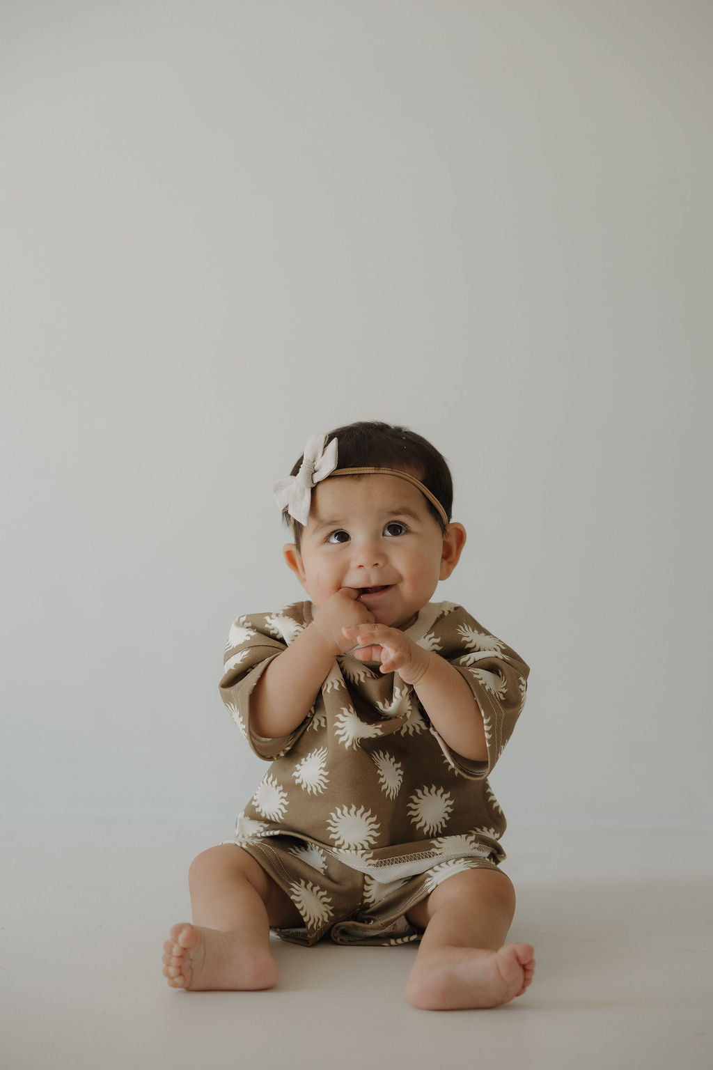 A baby wearing the forever french baby French Terry Short Set | Sol—brown short sleeves and matching shorts with white suns—plus a light bow headband, sits on the floor against a plain background, smiling and clapping hands.
