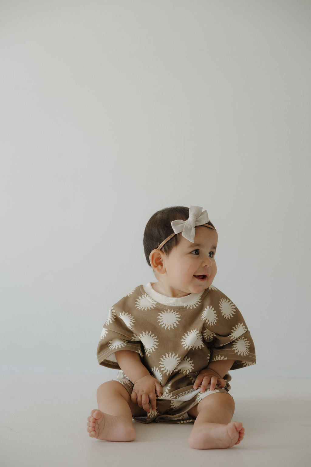 A baby with short dark hair and a light bow headband sits on the floor, smiling to the side in the forever french baby French Terry Short Set | Sol—an outfit featuring white sunburst patterns and matching shorts, against a plain light background.