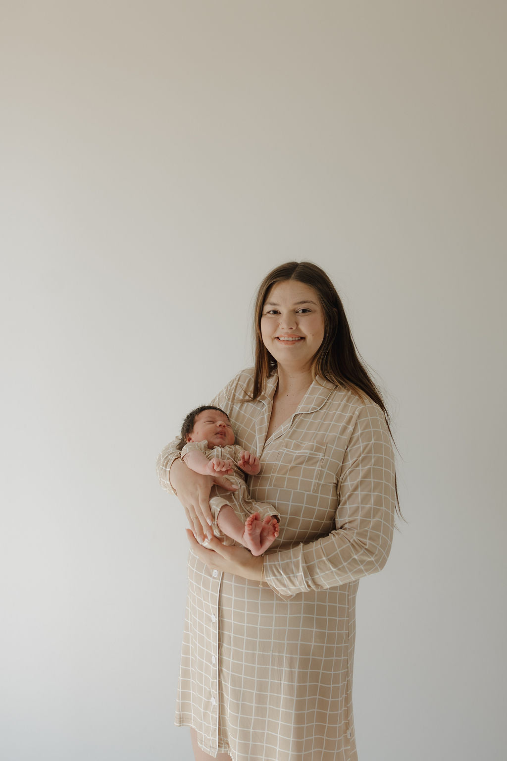 A woman with long brown hair smiles gently as she holds a newborn, wearing the forever french baby Women's Bamboo Sleeping Dress | Grid, against a plain light background.