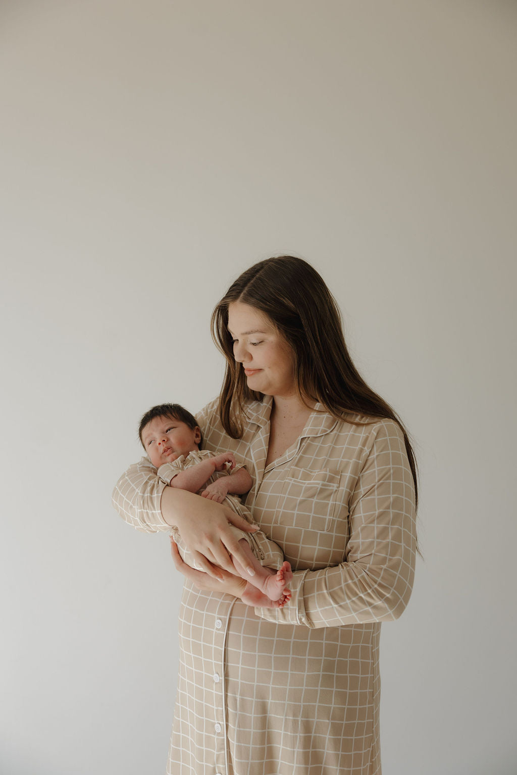 A woman with long brown hair wears the forever french baby Women's Bamboo Sleeping Dress | Grid in beige plaid as she gently smiles down at a newborn in her arms against a plain light background.