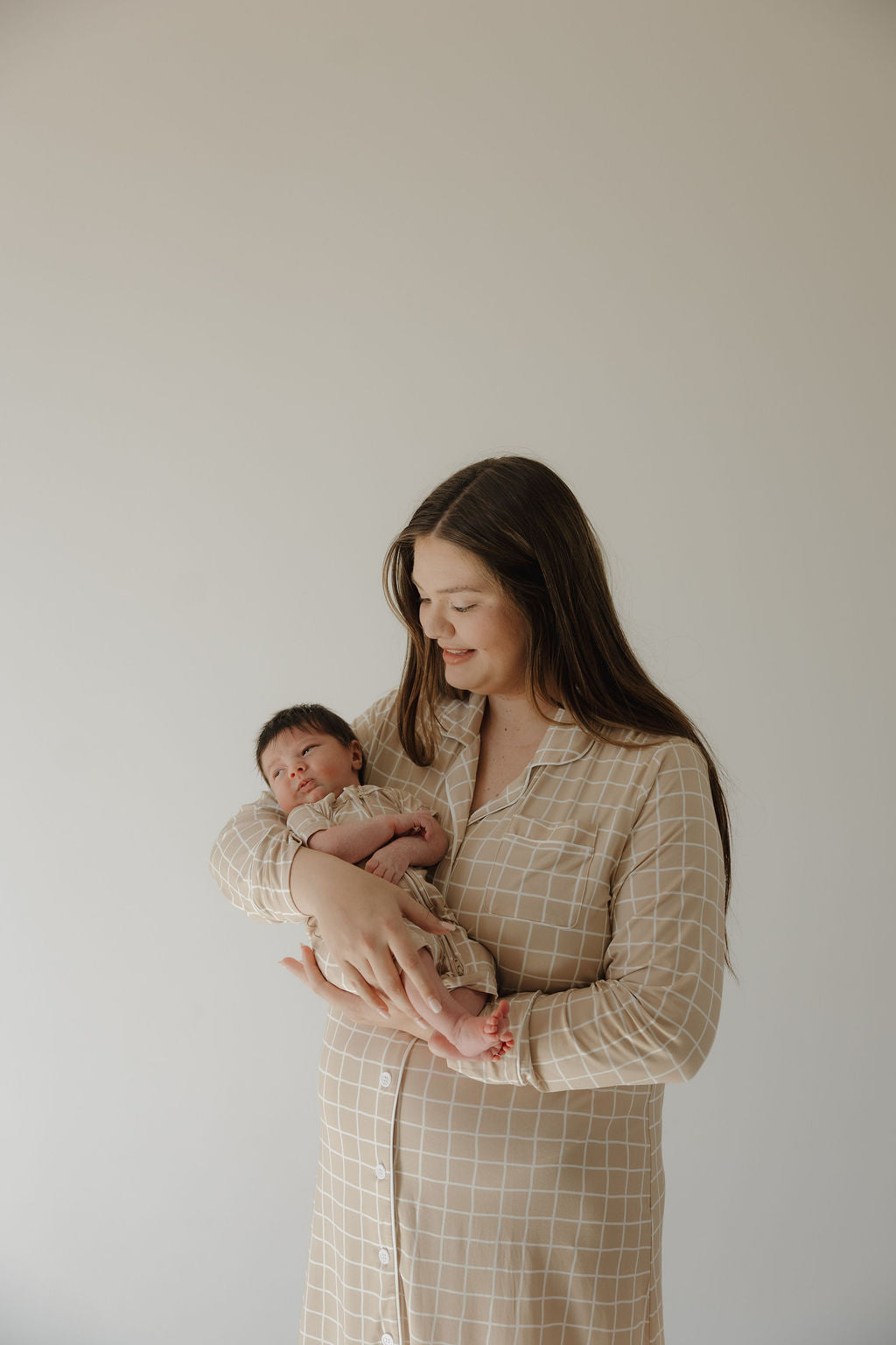 A woman with long brown hair, wearing the forever french baby Women's Bamboo Sleeping Dress | Grid, gently cradles a newborn and smiles softly against a plain light background.