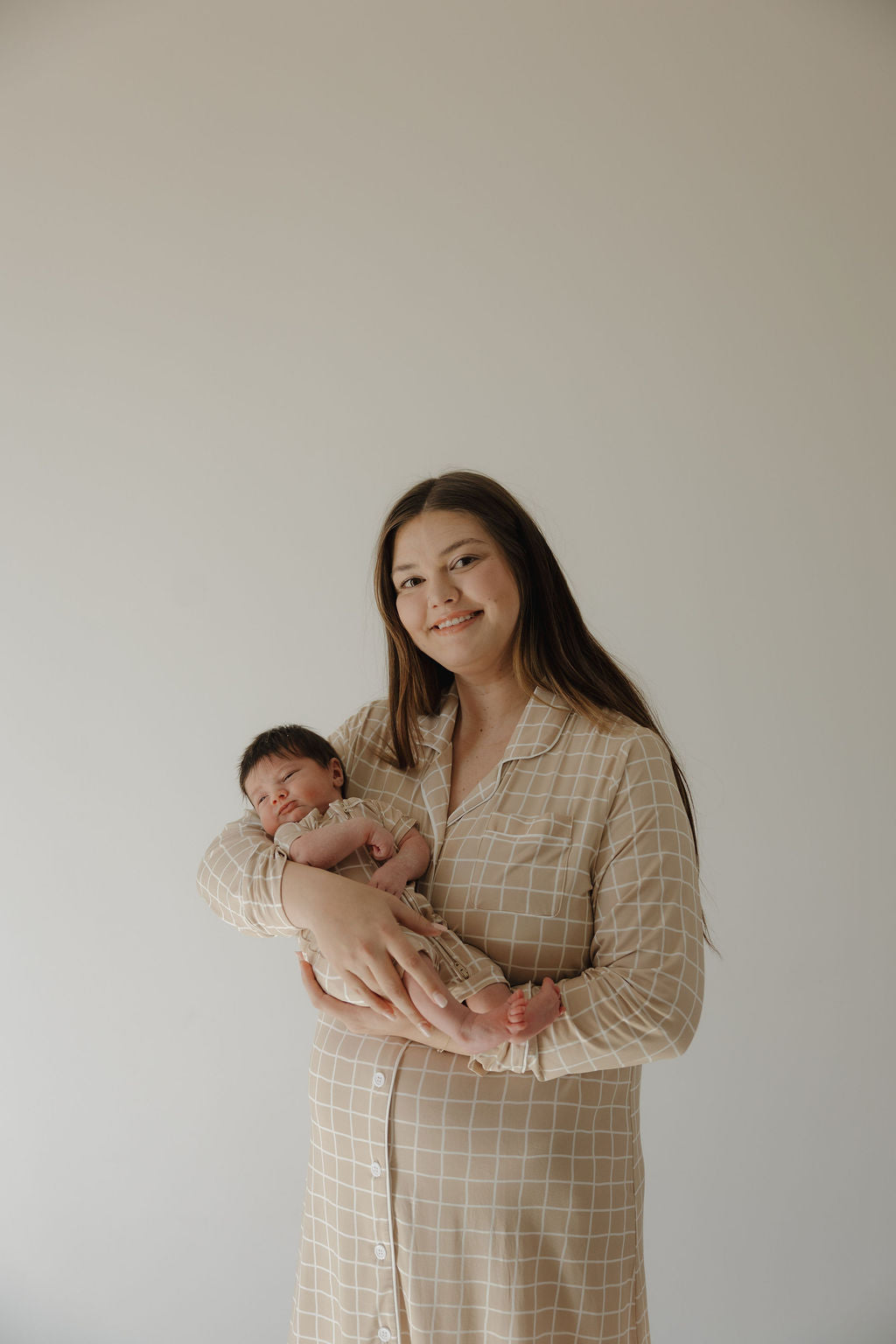 A woman with long brown hair smiles while holding a sleeping newborn, both wearing matching forever french baby Women's Bamboo Sleeping Dress | Grid outfits in soft, hypo-allergenic bamboo fabric, set against a plain, light background.