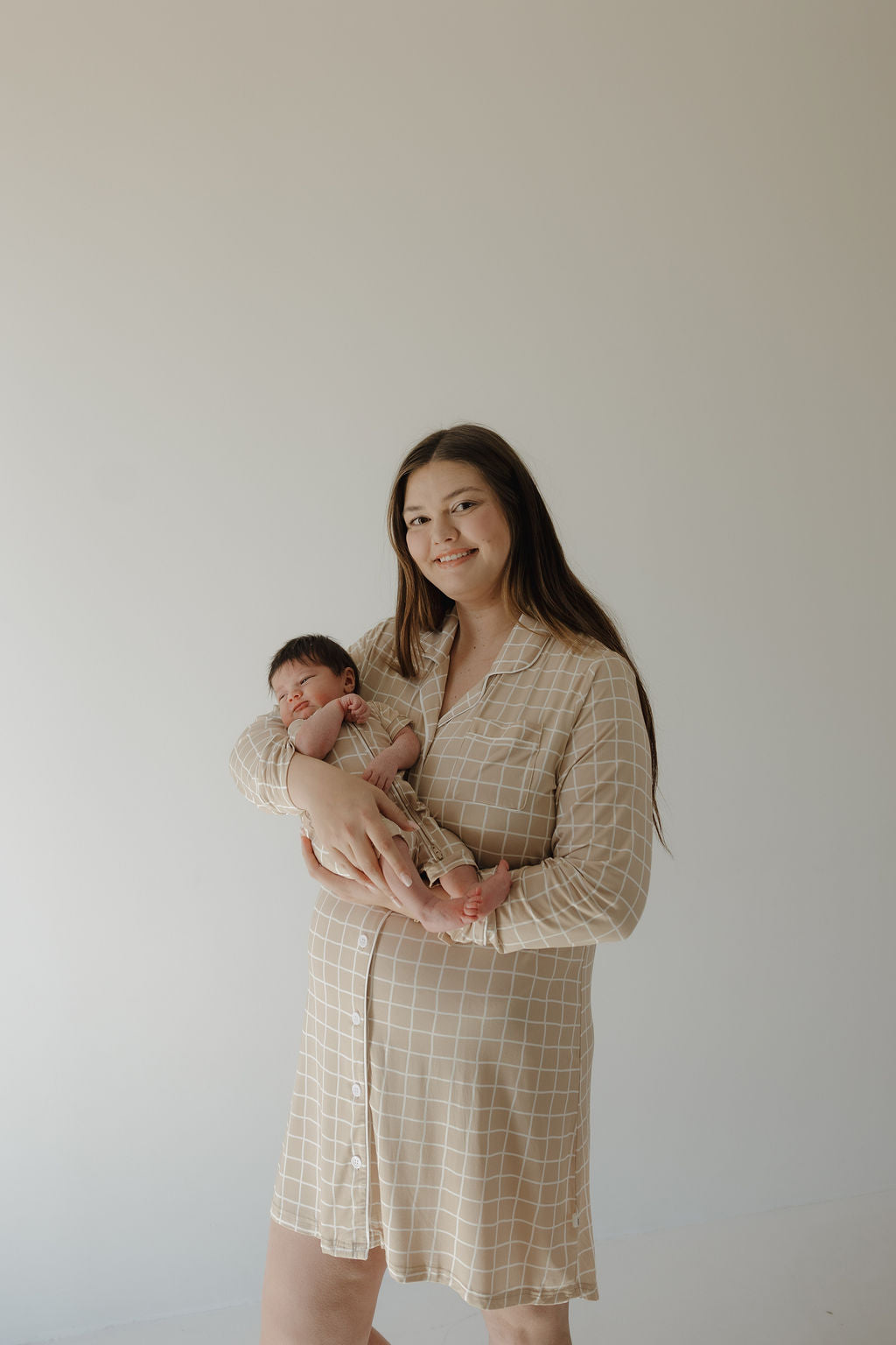 A woman wears the forever french baby Women's Bamboo Sleeping Dress | Grid and smiles gently against a plain background while holding a sleeping baby in a matching outfit.