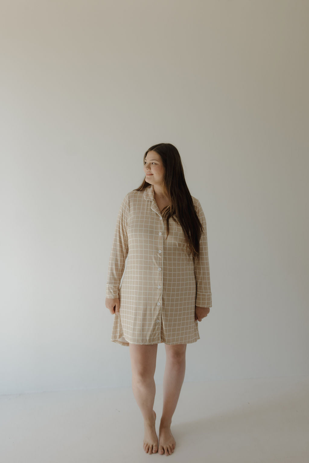 A woman with long brown hair stands barefoot against a plain backdrop, wearing the forever french baby Women's Bamboo Sleeping Dress | Grid, a loose beige plaid style made from hypo-allergenic bamboo fabric, and gazes to the side.