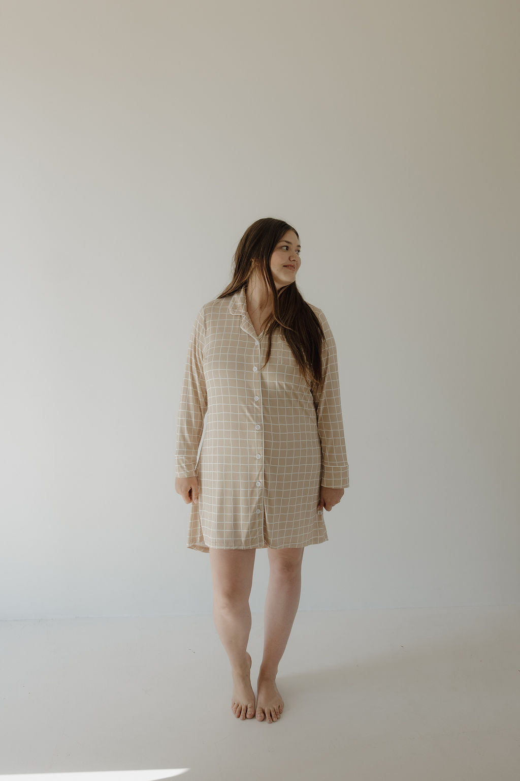 A woman with long brown hair stands barefoot in a minimal, light-filled room wearing the forever french baby Women's Bamboo Sleeping Dress | Grid, a beige plaid, long-sleeved sleep dress made from hypo-allergenic bamboo fabric, smiling as she looks to her left.
