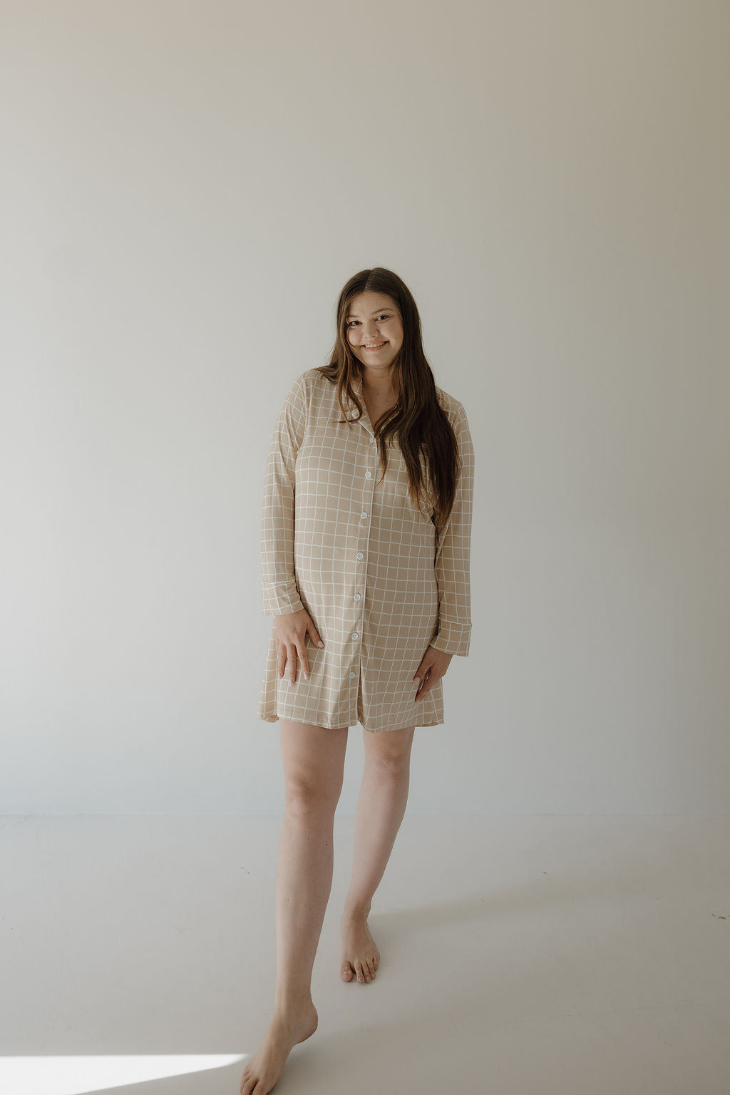 A woman with long brown hair stands barefoot on a white floor, wearing the Women's Bamboo Sleeping Dress | Grid by forever french baby, made from soft hypo-allergenic bamboo fabric, against a plain light background.