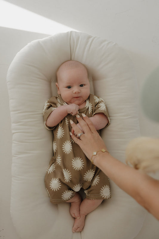 A baby in the forever french baby French Terry Romper | Sol, featuring white sun patterns, lies on a cushioned white lounger. An adult hand rests gently on the baby’s chest, with soft natural light highlighting the cozy outfit.