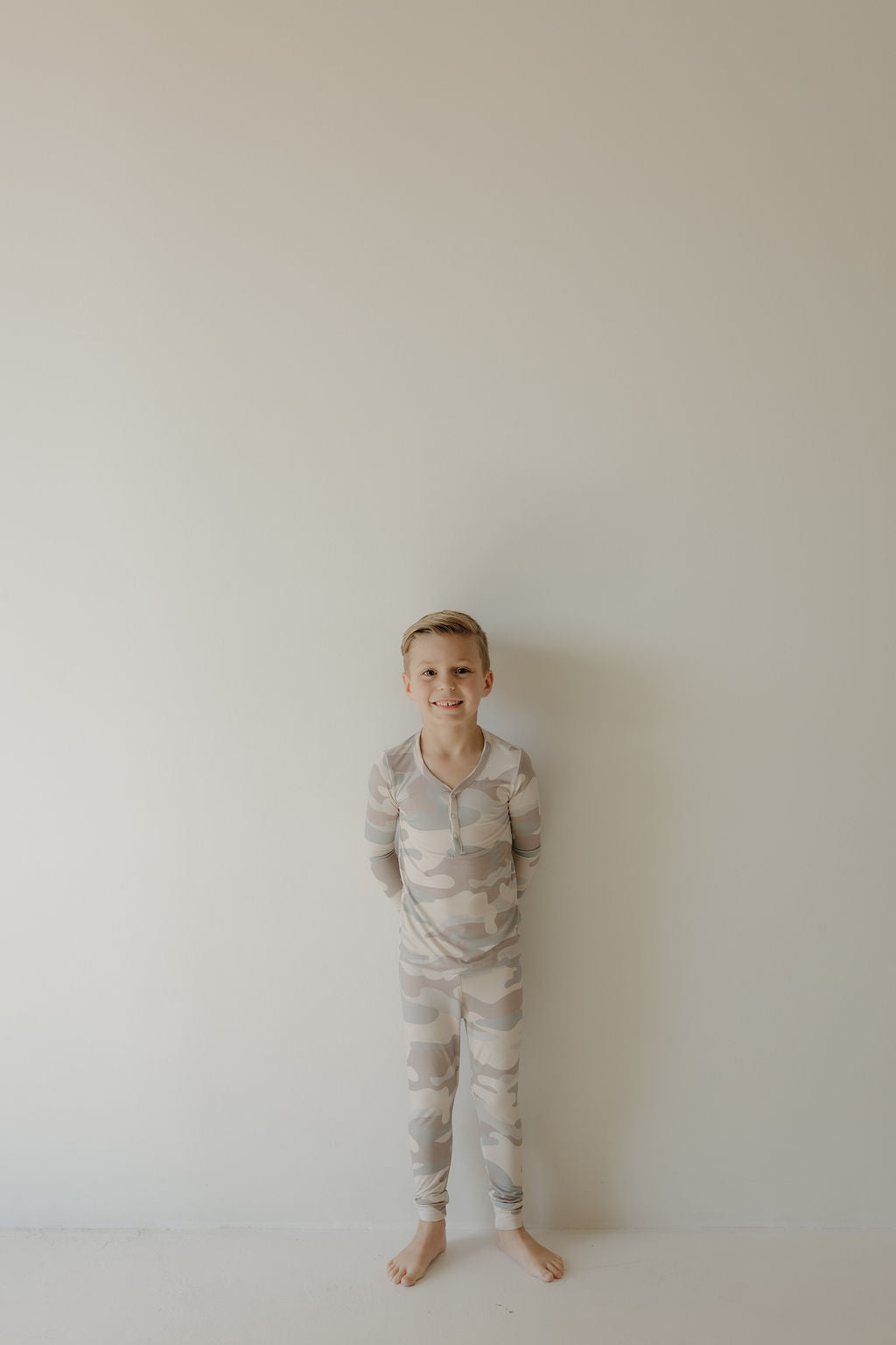 A young child with short hair stands barefoot by a plain wall, smiling in forever french baby's Bamboo Two Piece Pajamas | Camo, made from breathable bamboo fabric.