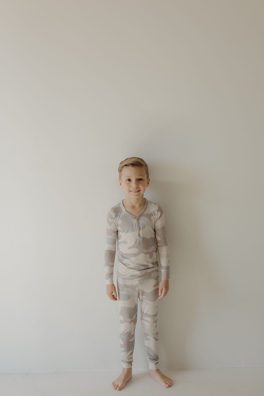 A smiling young boy with short blond hair stands barefoot against a plain wall, wearing forever french baby’s Bamboo Two Piece Pajamas | Camo, made from breathable bamboo fabric.