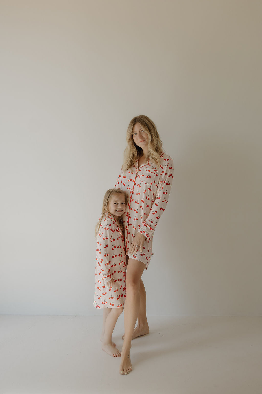 A woman and a young girl smile and pose barefoot in matching forever french baby pajamas, with the girl wearing the Toddler Bamboo Sleeping Dress | Cherry Orchard, featuring a red heart pattern on soft bamboo fabric against a light background.