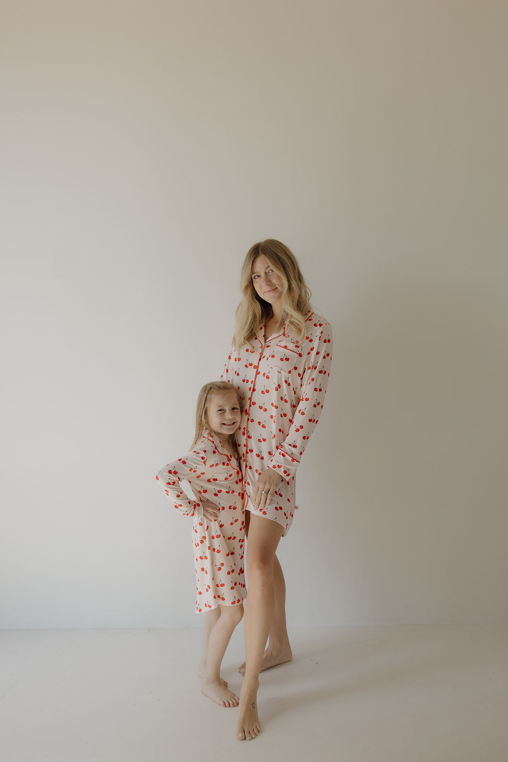 A woman and young girl stand barefoot against a plain wall, both wearing forever french baby Toddler Bamboo Sleeping Dresses in the Cherry Orchard print. The girl smiles with her hand on her hip as the woman stands beside her, smiling softly.