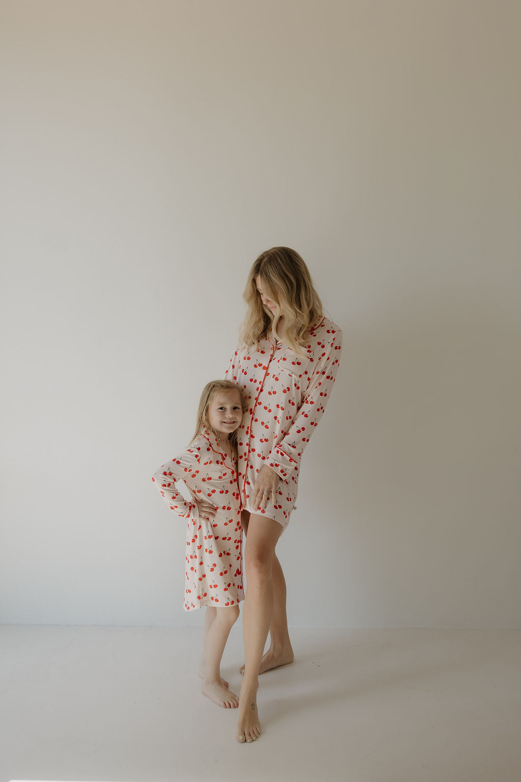 A woman and a young girl stand barefoot, both wearing forever french baby’s Toddler Bamboo Sleeping Dress | Cherry Orchard. Made from hypo-allergenic bamboo with cherry patterns, the soft dress ensures comfort as the woman gently hugs the girl.