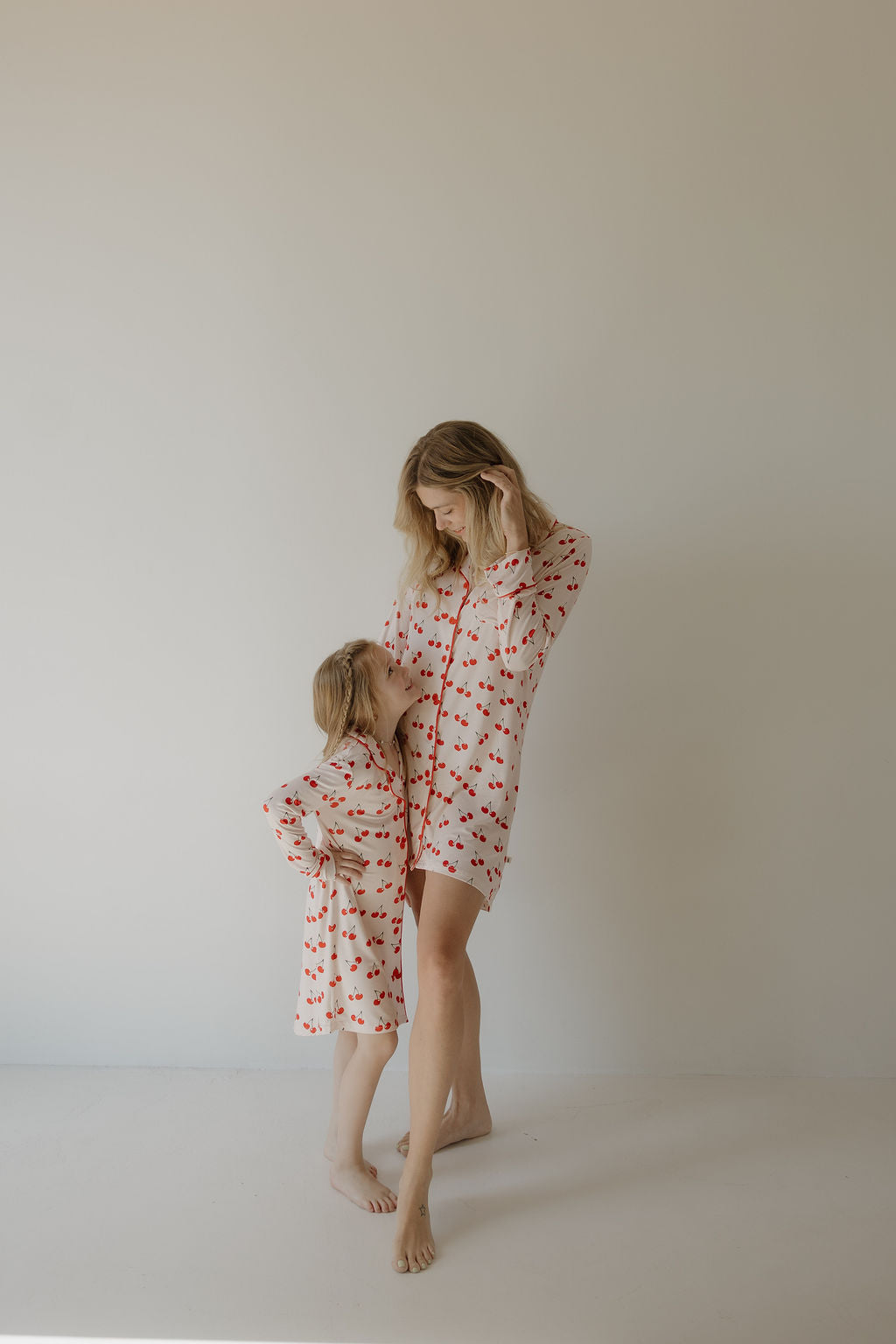 A woman and young girl stand barefoot by a light wall, both in matching forever french baby bamboo outfits. The girl, in her cozy Toddler Bamboo Sleeping Dress | Cherry Orchard, leans affectionately against the woman.
