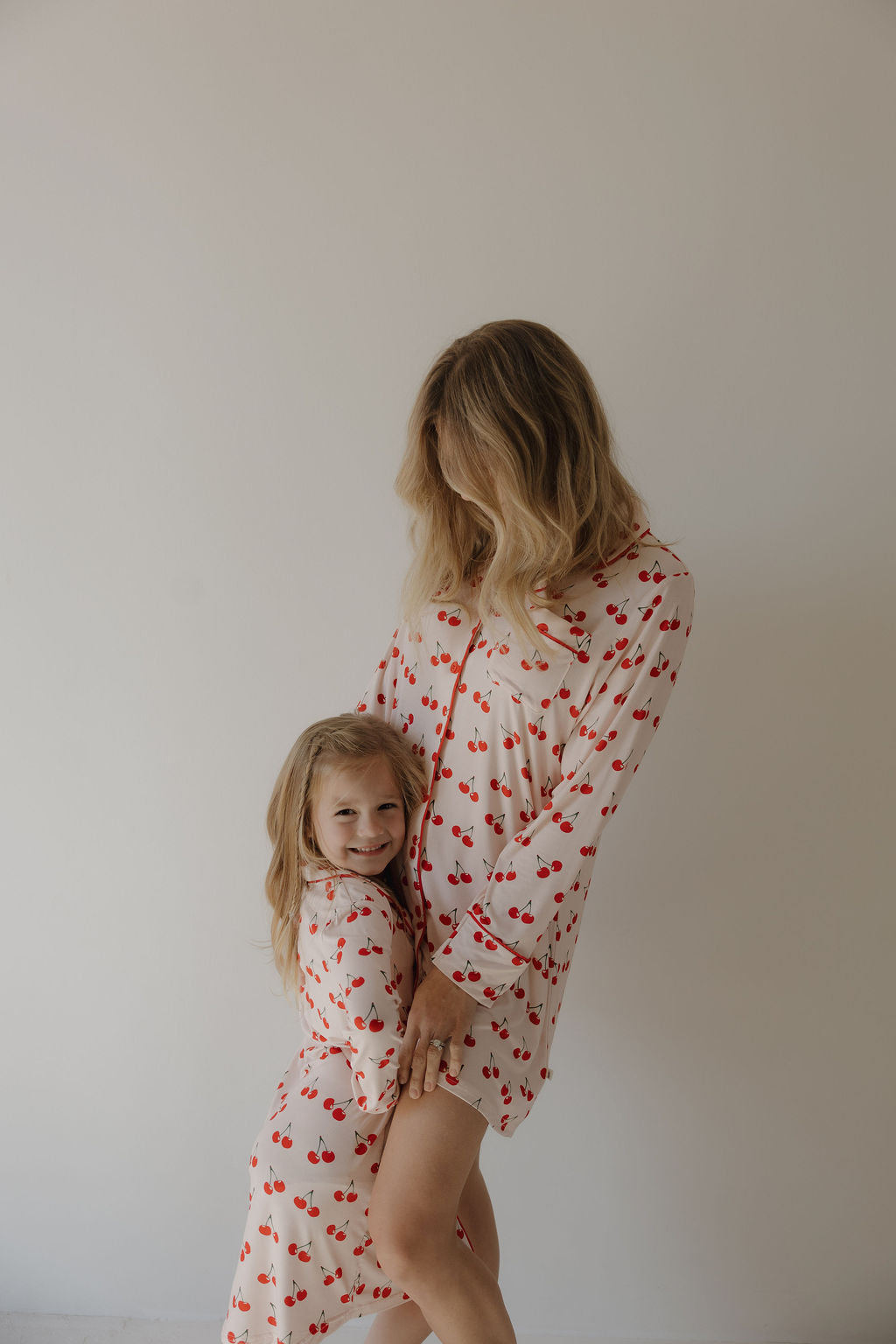 A woman and a young girl pose together in matching forever french baby cream-colored pajamas with red cherry prints. The woman wears the Women's Bamboo Sleeping Dress | Cherry Orchard as the girl smiles and hugs her leg against a plain background.