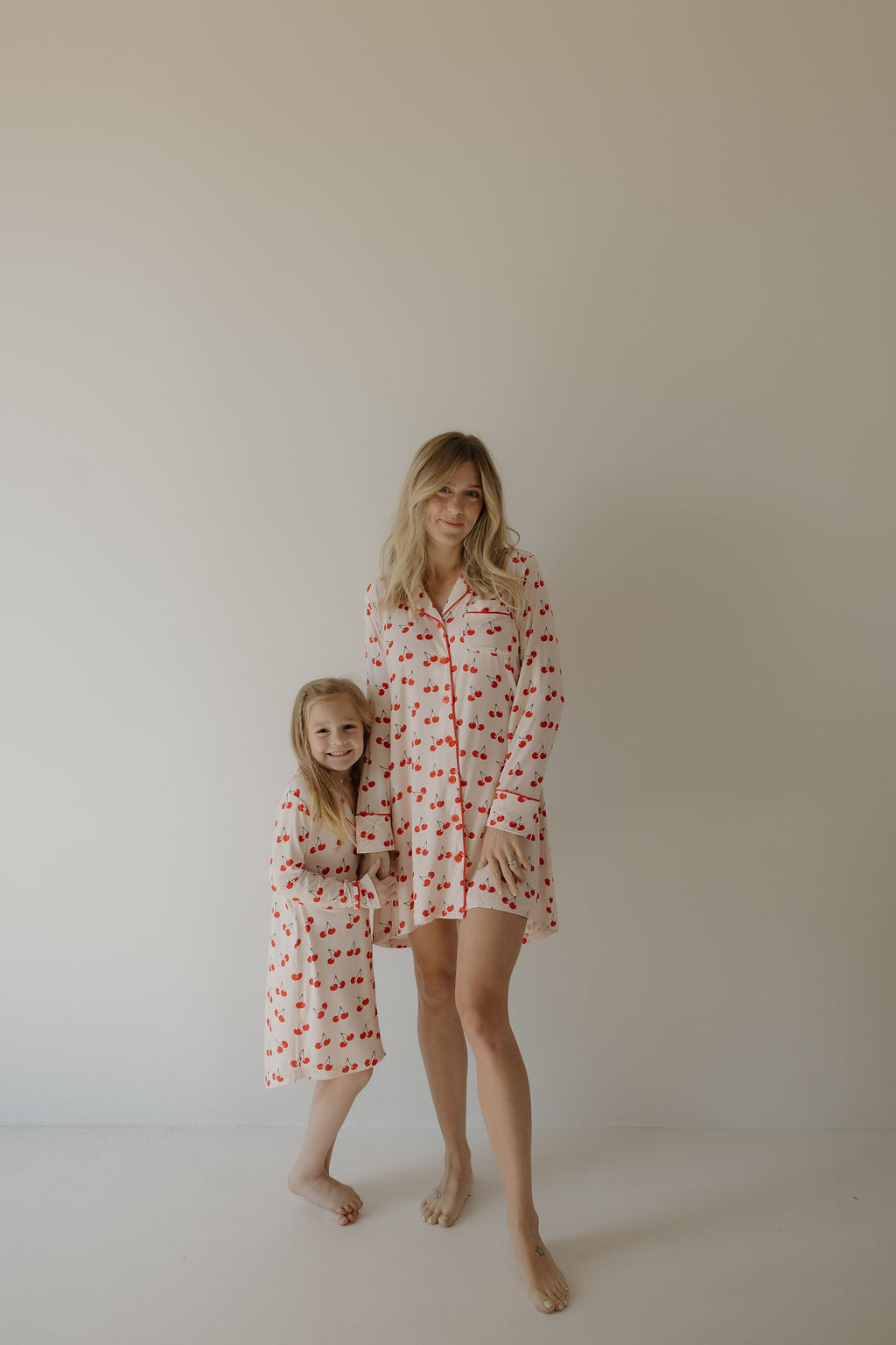 A woman and a young girl, both barefoot and smiling, wear matching forever french baby Women's Bamboo Sleeping Dresses in the Cherry Orchard print featuring red hearts, posing together against a plain light background.