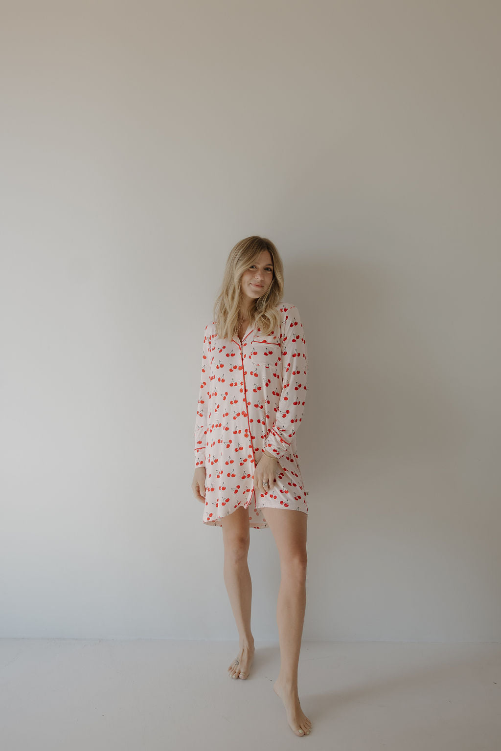 A woman with long blonde hair stands barefoot against a light wall, wearing the forever french baby Women's Bamboo Sleeping Dress | Cherry Orchard, featuring a white fabric with red cherry print and red trim.