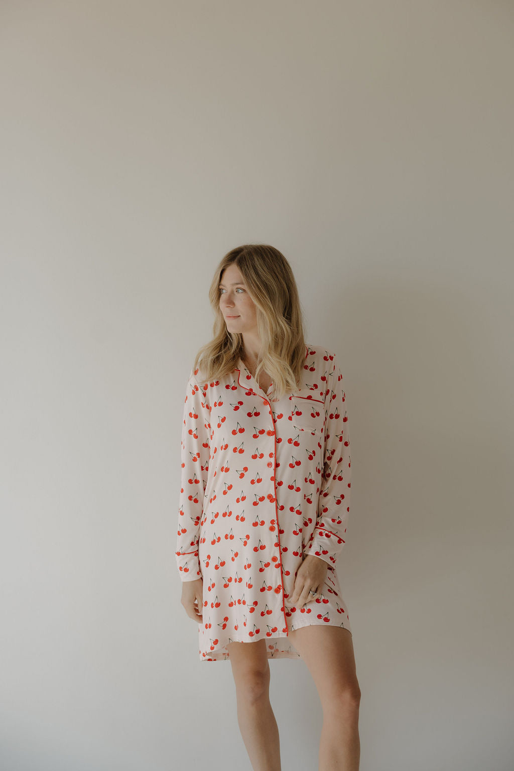 A woman with long blonde hair models the forever french baby Women's Bamboo Sleeping Dress | Cherry Orchard, featuring a hypo-allergenic cherry pattern, against a plain light background while looking to the side.