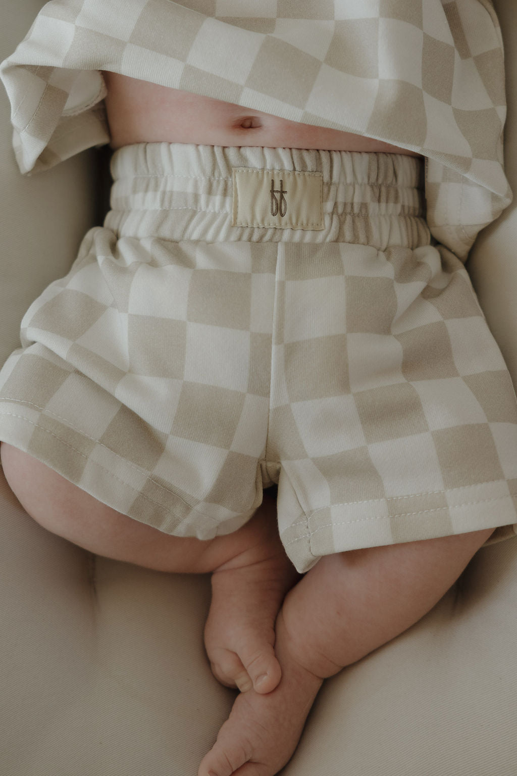 A baby wearing the forever french baby French Terry Short Set in a beige and white checkerboard pattern, features a soft short sleeve top and matching shorts with an FF logo, lying down with legs slightly bent and feet crossed.