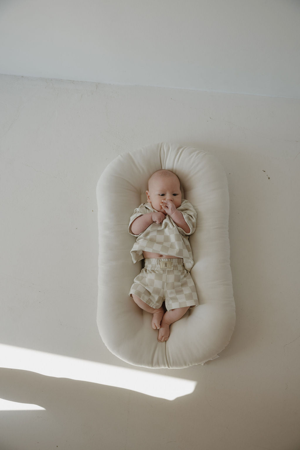 A baby lies on a cream cushion wearing the forever french baby French Terry Short Set | Checkerboard, a beige and white checkered short sleeve top with matching shorts. Sunlight streams in as the baby rests on a light-colored floor.