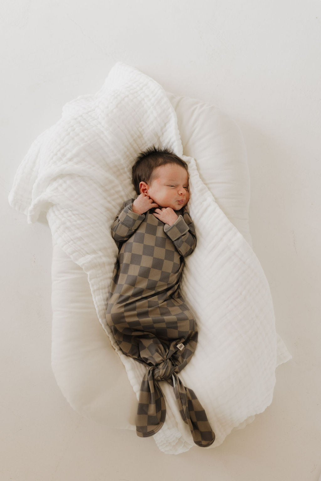 A newborn with dark hair, wearing a forever french baby Bamboo Knotted Gown in Faded Checkerboard, peacefully sleeps on a soft, white cushion. The hypo-allergenic outfit provides comfort as the baby rests on its back with hands gently curled near its face.