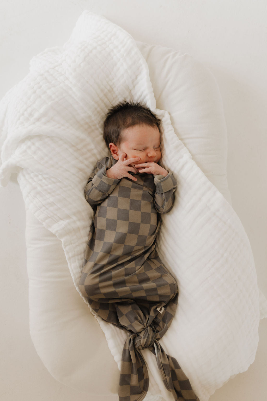 A newborn sleeps peacefully on a soft, white blanket, wearing the forever french baby Bamboo Knotted Gown in Faded Checkerboard. The brown and gray checkered outfit is a newborn essential that enhances the cozy setup with warmth and tranquility.