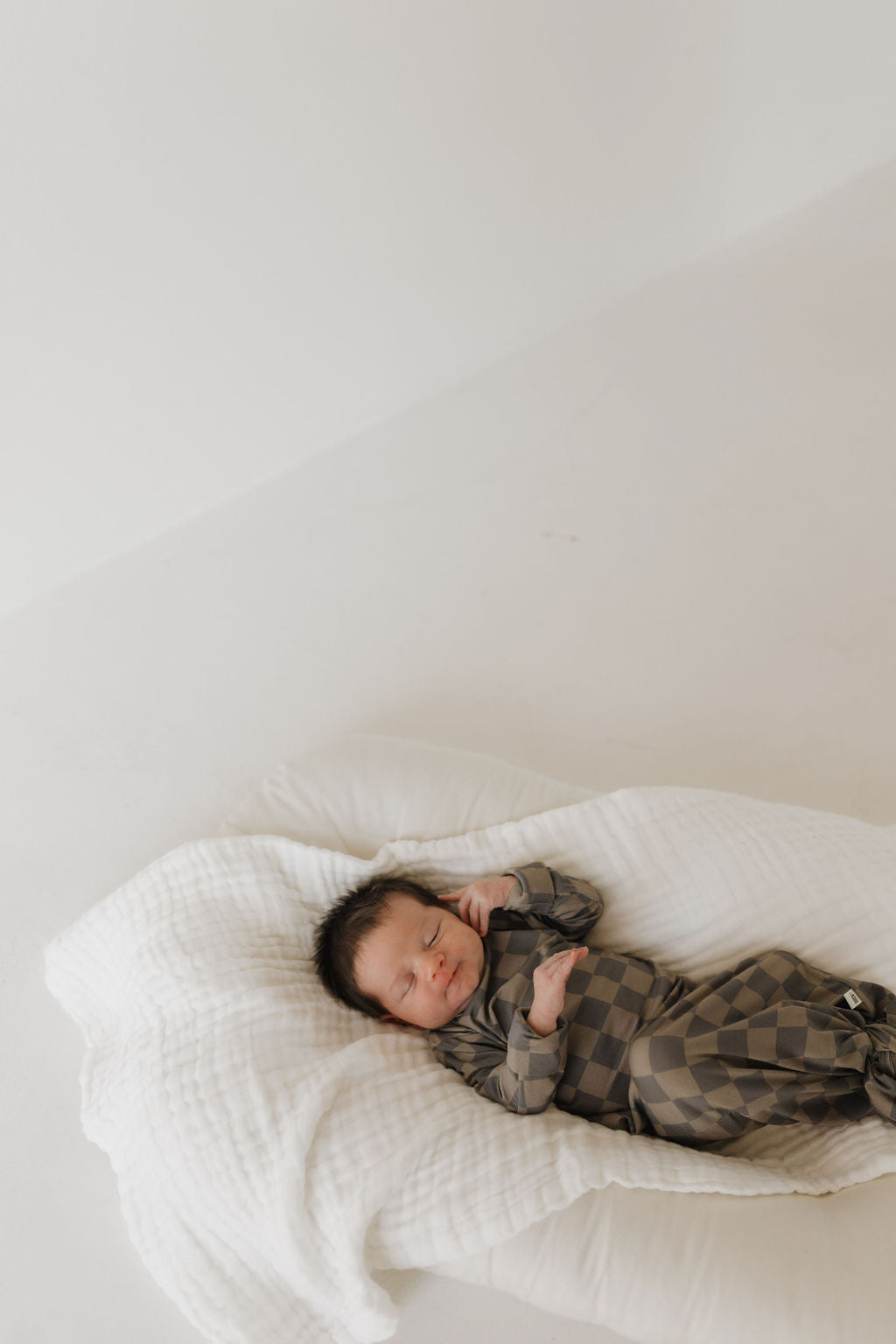 A newborn in a cozy Bamboo Knotted Gown, Faded Checkerboard by forever french baby is peacefully sleeping on a soft white blanket. The minimalistic setting with a light, neutral background is perfect for easy diaper changes.