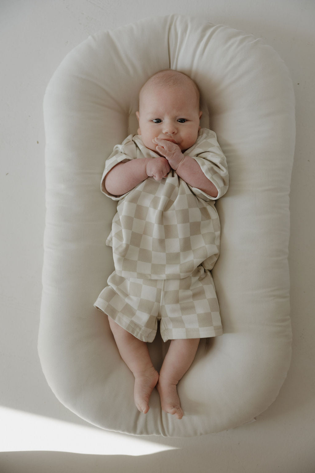 A baby wearing the forever french baby French Terry Short Set | Checkerboard in beige and white lies on a soft, cream cushion, gazing upward with hands by their mouth and feet stretched out.